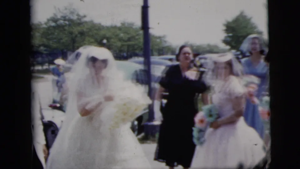 The veiled bride carries a light bouquet while bridesmaids and an adult guest walk beside parked mid-century cars.