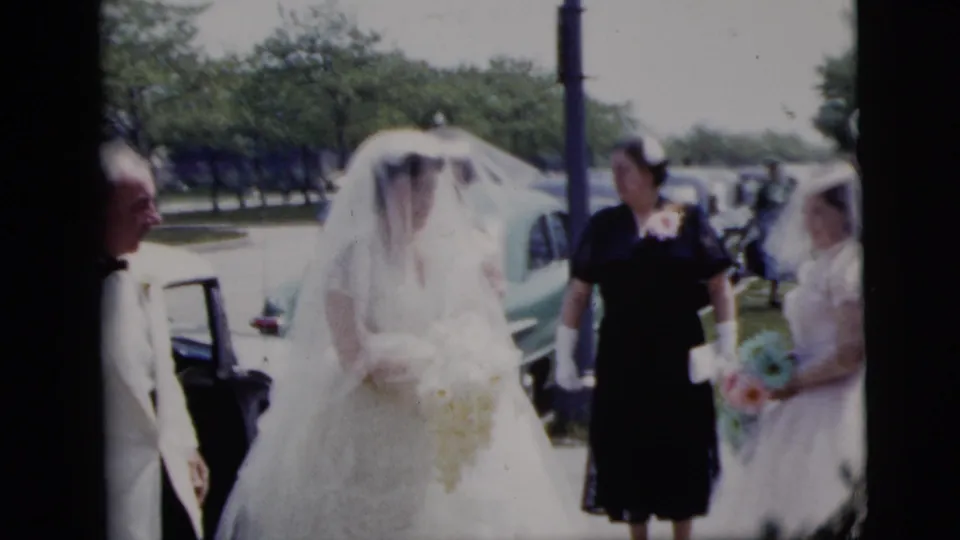 A veiled bride in a white gown appears near bridesmaids with bouquets and adult guests on a sunlit New York street.