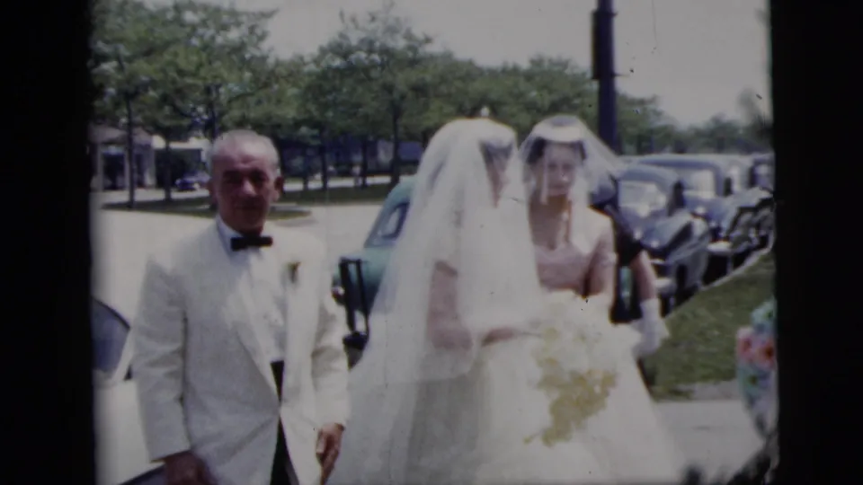 A man in a white tuxedo accompanies the veiled bride as another attendant follows near parked cars and trees.