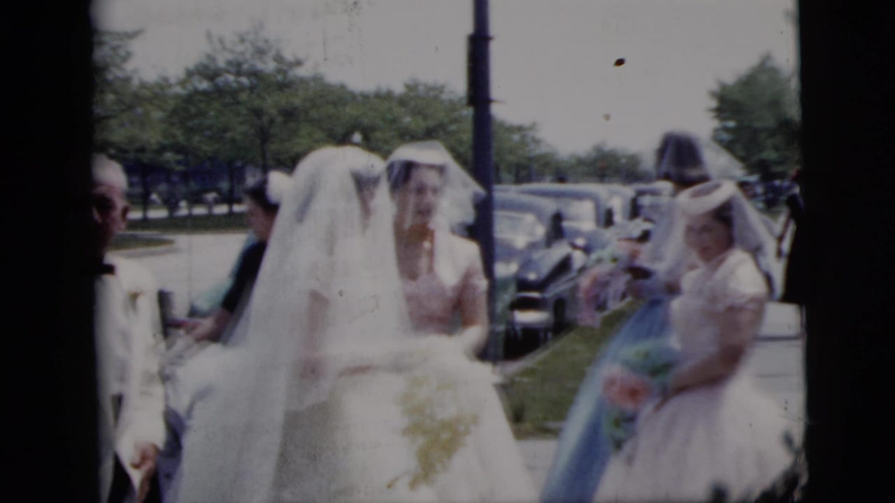 In 1958 New York City home-movie footage, brides in white gowns and veils walk outdoors with attendants near parked mid-century cars.