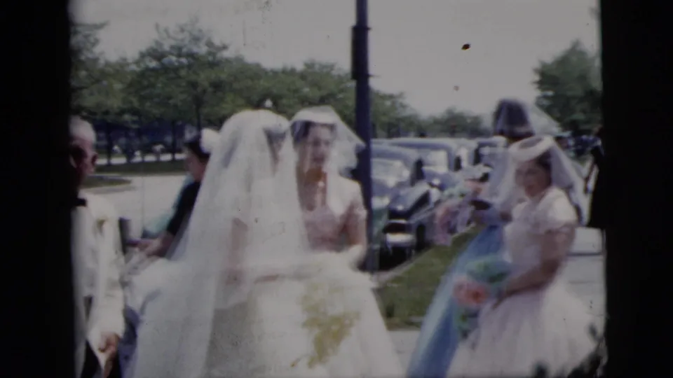 In 1958 New York City home-movie footage, brides in white gowns and veils walk outdoors with attendants near parked mid-century cars.