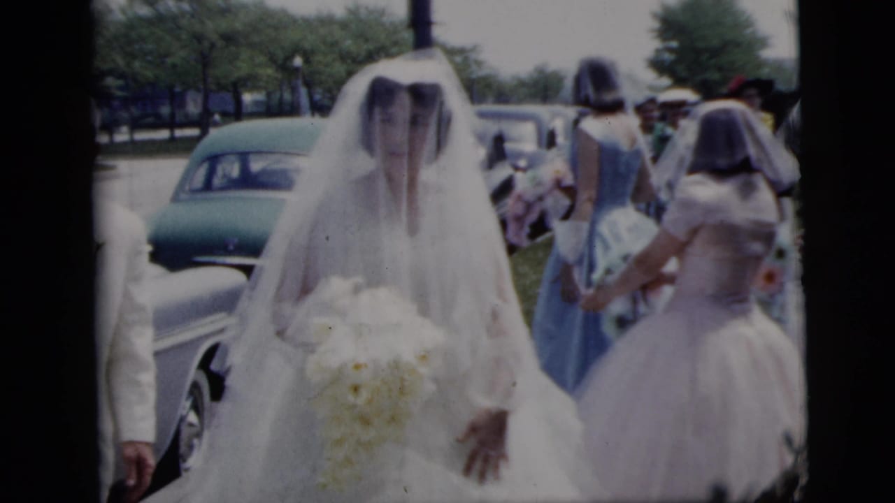 A veiled bride carrying a bouquet walks near bridesmaids and parked cars.