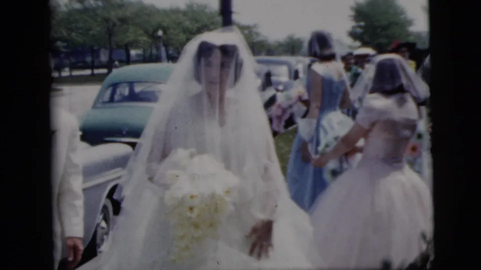 A veiled bride with a white bouquet walks outdoors in 1958 New York City, with bridesmaids and vintage cars behind her.