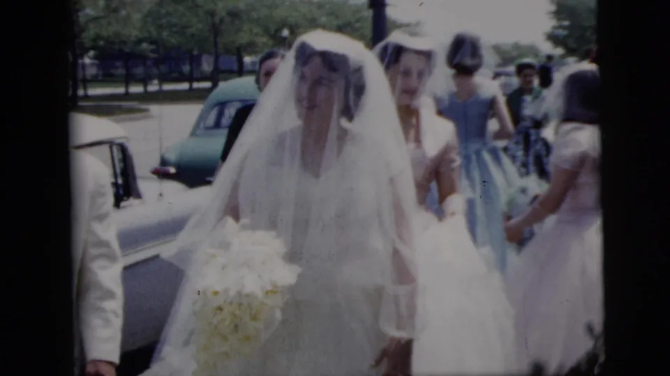 A bride in a long white veil approaches the home-movie camera near a parked 1950s automobile, with attendants visible behind her.