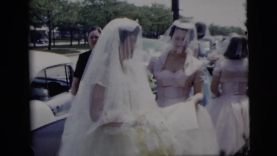 A veiled bride and a bridesmaid in a pink dress walk together beside an open car door during the outdoor wedding scene.