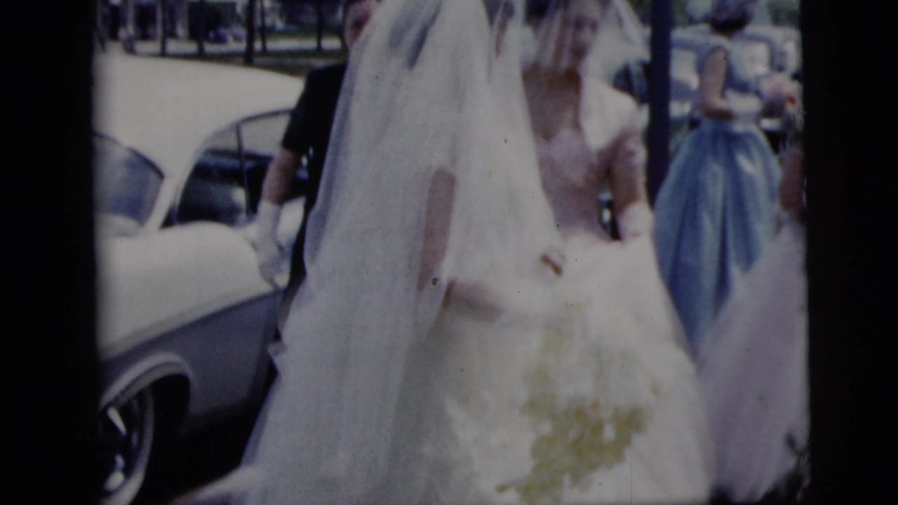 In supported 1958 New York City home-movie footage, a veiled bride in a white gown stands near a white car with attendants behind her.