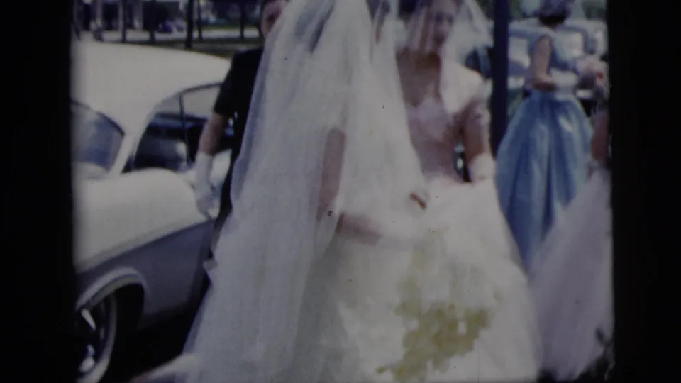 In supported 1958 New York City home-movie footage, a veiled bride in a white gown stands near a white car with attendants behind her.