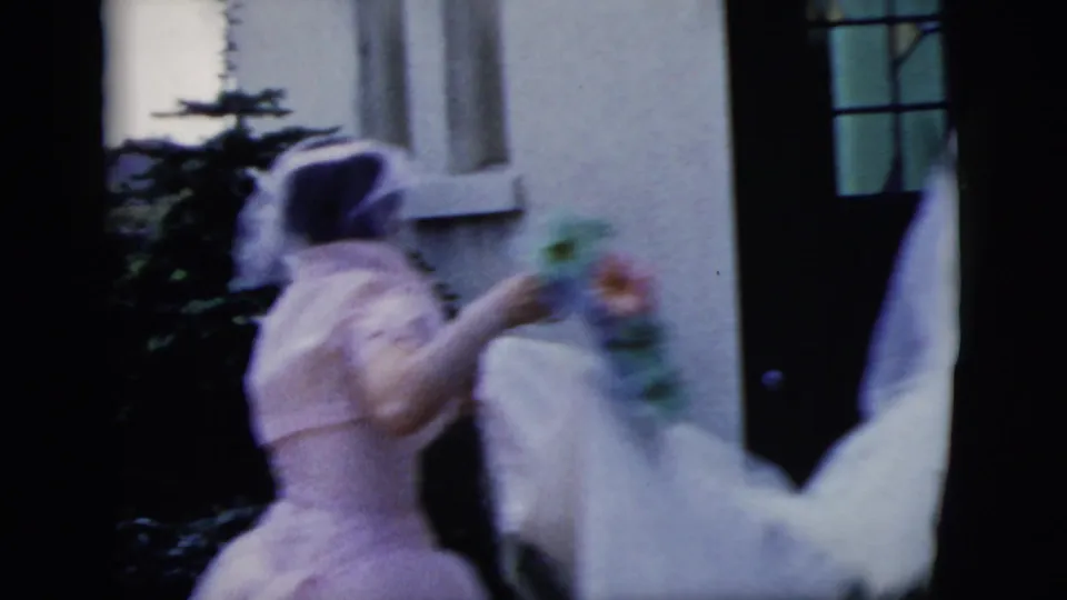 The veiled bride, holding a bouquet, moves close to the church entrance while a dark-suited figure passes in the foreground.