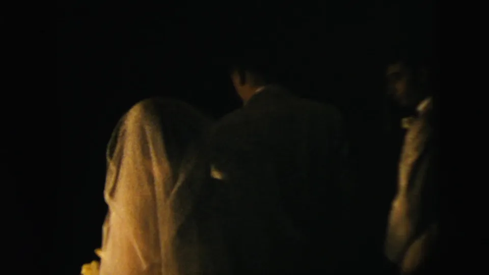 Low-light wedding ceremony frame showing the bride and groom from behind with another suited man nearby.