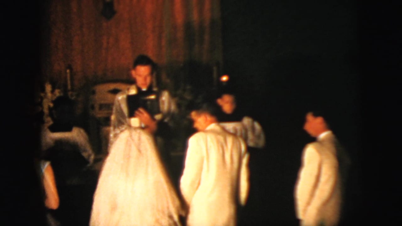 Bride and groom kneel before clergy during a dimly lit church wedding ceremony.