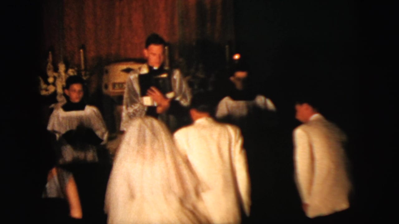 1958 New York City wedding frame with the veiled bride and groom before an officiant, surrounded by attendants and clergy in a dark church interior.