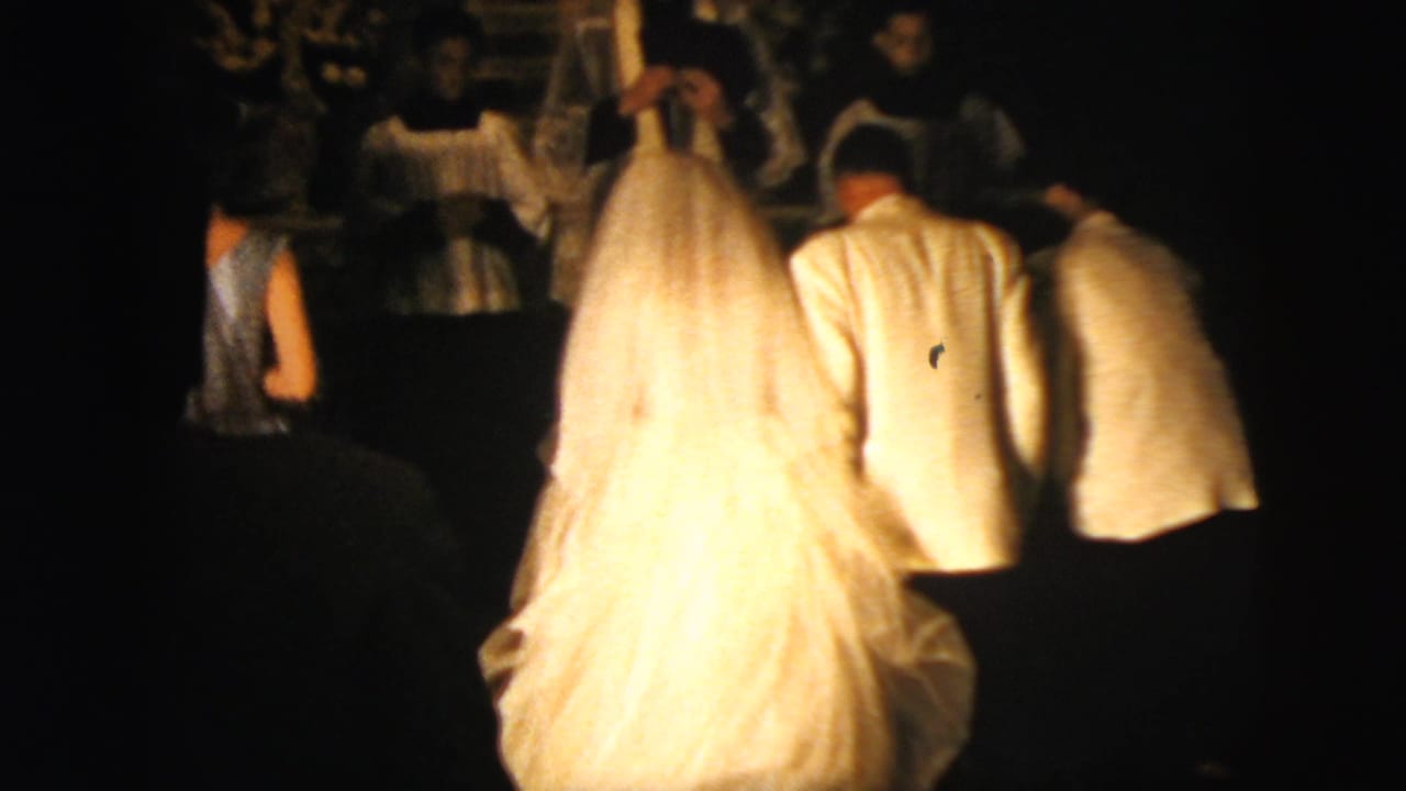 In 1958 New York City home-movie footage, a bride in a white gown and veil moves through a dim church aisle with formal attendants around her.