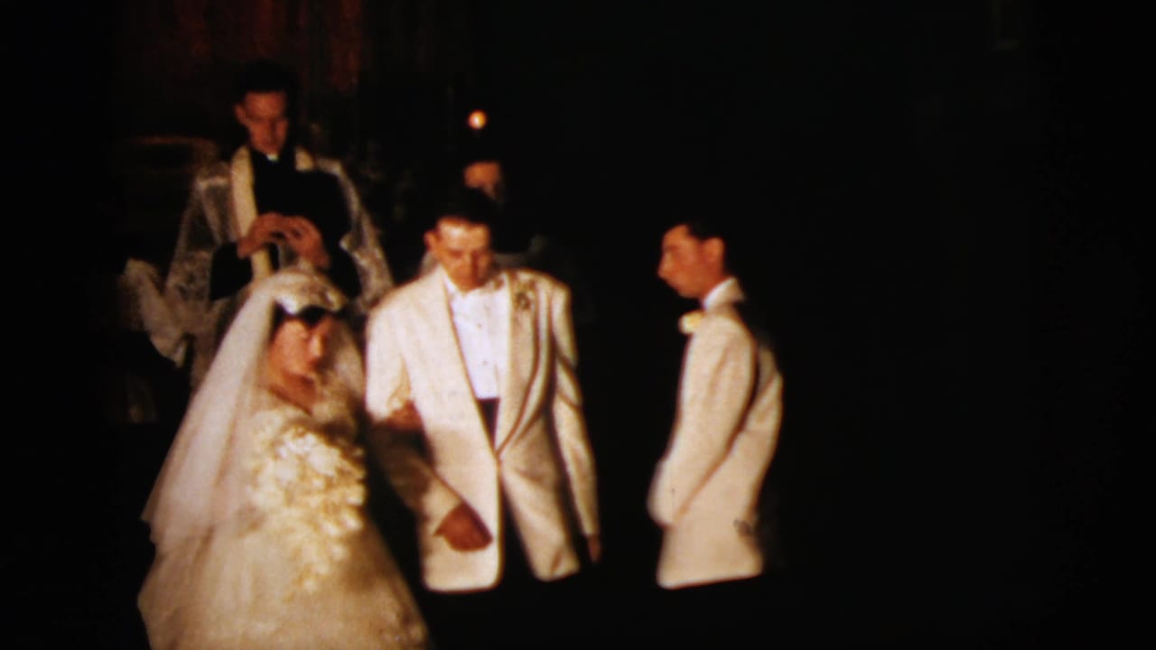 Bride and groom in formal wedding attire stand arm-in-arm near white-vested clergy or attendants inside a dim New York City church.