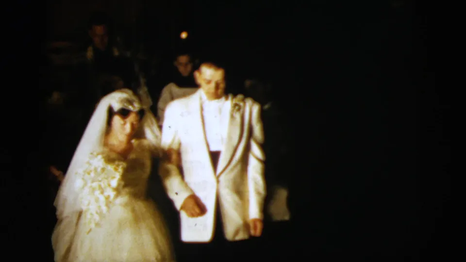 Bride and groom kneel in the shadowed church interior, with the groom's white jacket and the bride's veil catching the strongest light.