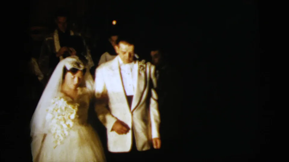 1958 New York City home-movie frame of a bride in veil and groom in light tuxedo walking arm-in-arm at night.