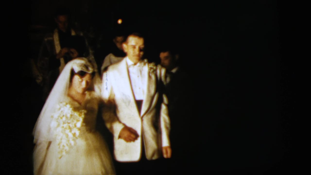 The bride and groom remain arm-in-arm in a dark nighttime wedding scene, with faces softly lit and guests behind them.