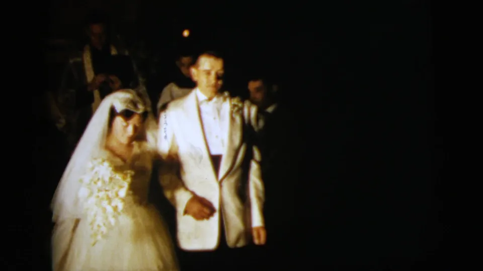 The bride and groom remain arm-in-arm in a dark nighttime wedding scene, with faces softly lit and guests behind them.