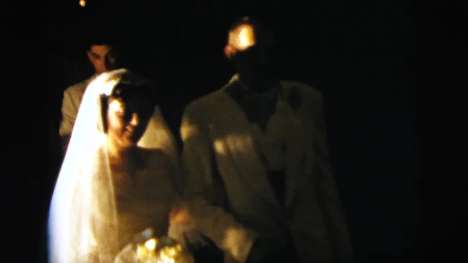 Bride and groom move toward the handheld 8mm camera, with faces softly lit in a dark wedding setting.