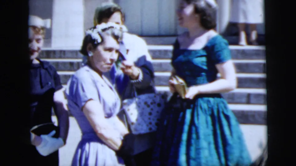 Women in mid-century dresses stand on exterior steps, likely among wedding guests in the New York City home movie.