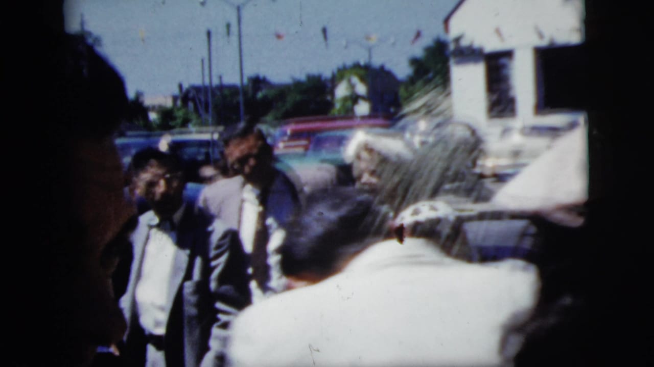 Wedding guests gather on a sunny street near parked cars and houses.