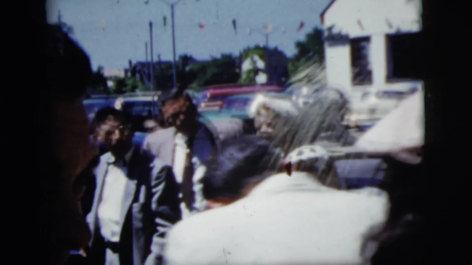 1958 New York City home-movie frame showing wedding guests walking outside near parked cars, houses, and trees.