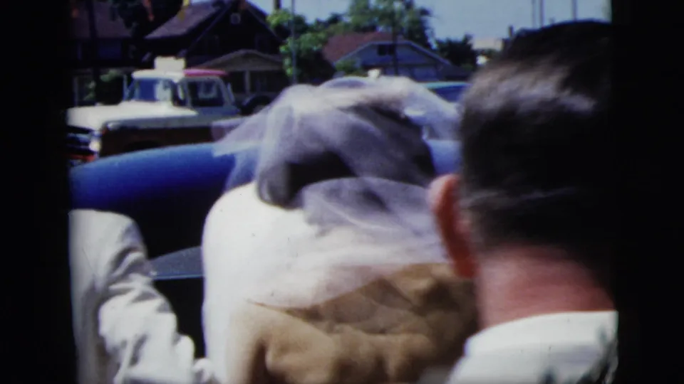 1958 New York City wedding home-movie frame with the bride's veil, guests, a blue car, and a white truck in the background.