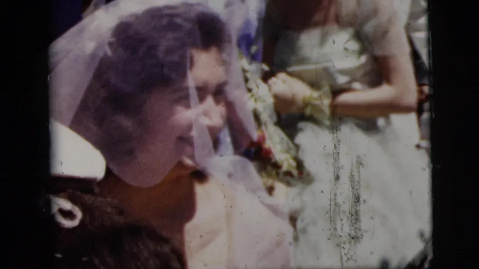 1958 New York City wedding frame showing the bride smiling beside a woman with flowers amid the outdoor group.