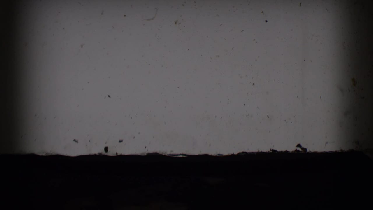Low-light 8mm frame from the Saint Louis clip showing a pale interior wall or surface, dark foreground band, and visible film artifacts.