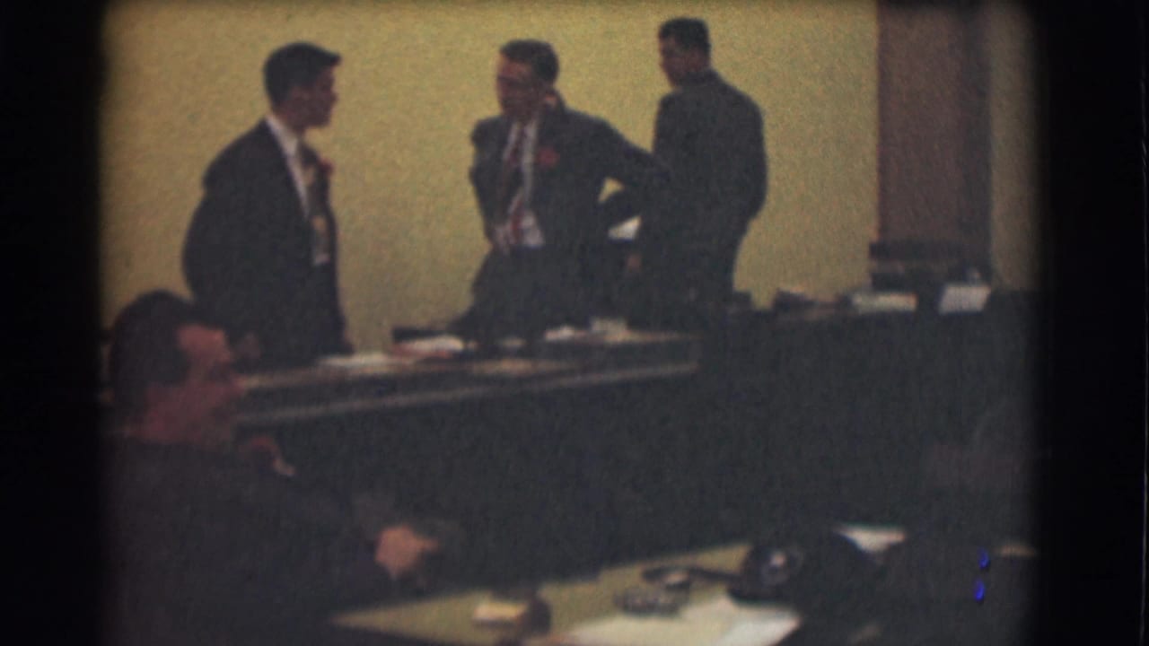 Saint Louis 1959 office scene with three men in suits standing by a long desk and a seated man partly visible in the foreground.