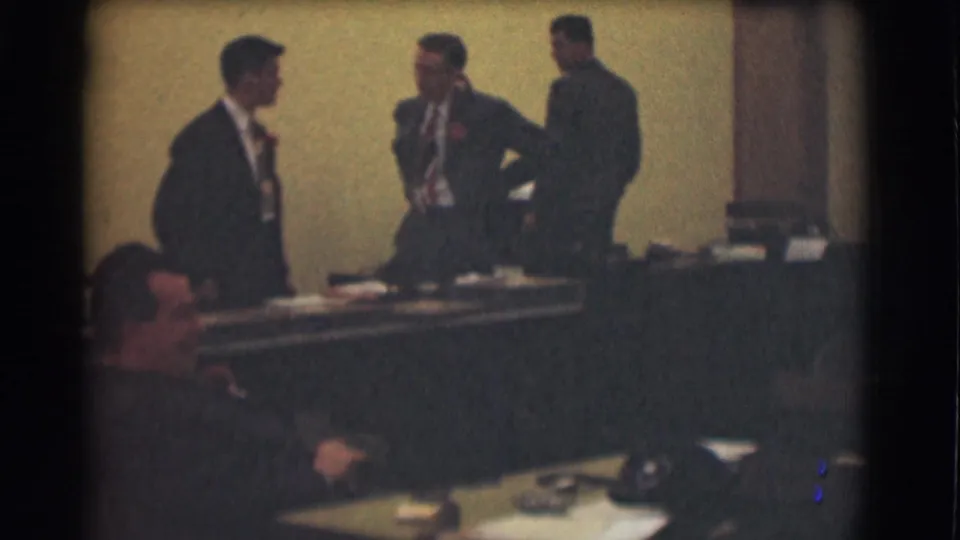 Saint Louis 1959 office scene with three men in suits standing by a long desk and a seated man partly visible in the foreground.