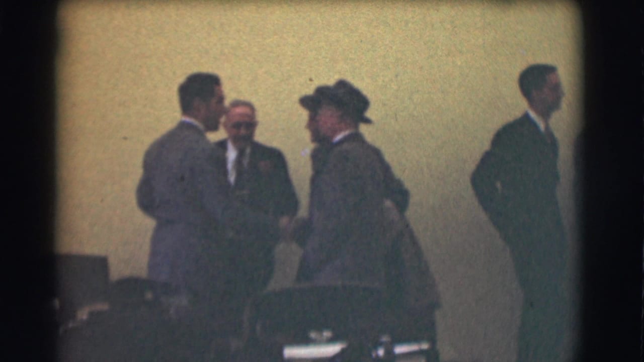 Four suited men stand and talk in a dim interior meeting room.