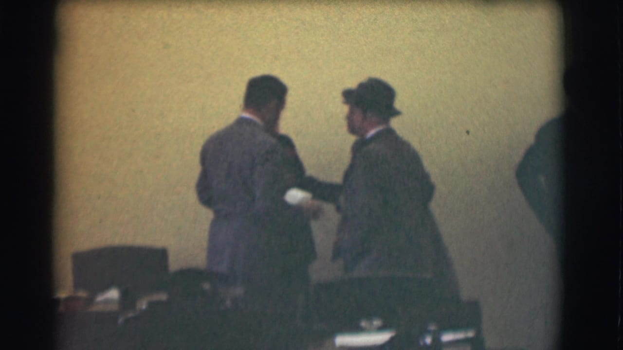 A suited man and a hat-wearing man converse in a 1959 Saint Louis interior, with chairs and a pale wall behind them.