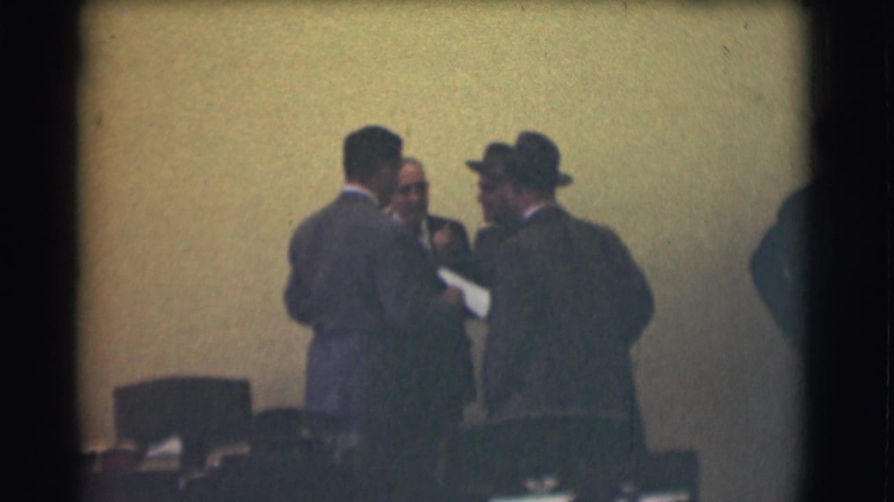 Three adult men in suits appear to confer in a Saint Louis interior, with chairs and meeting-room objects in the foreground.