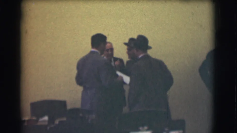 Three adult men in suits appear to confer in a Saint Louis interior, with chairs and meeting-room objects in the foreground.