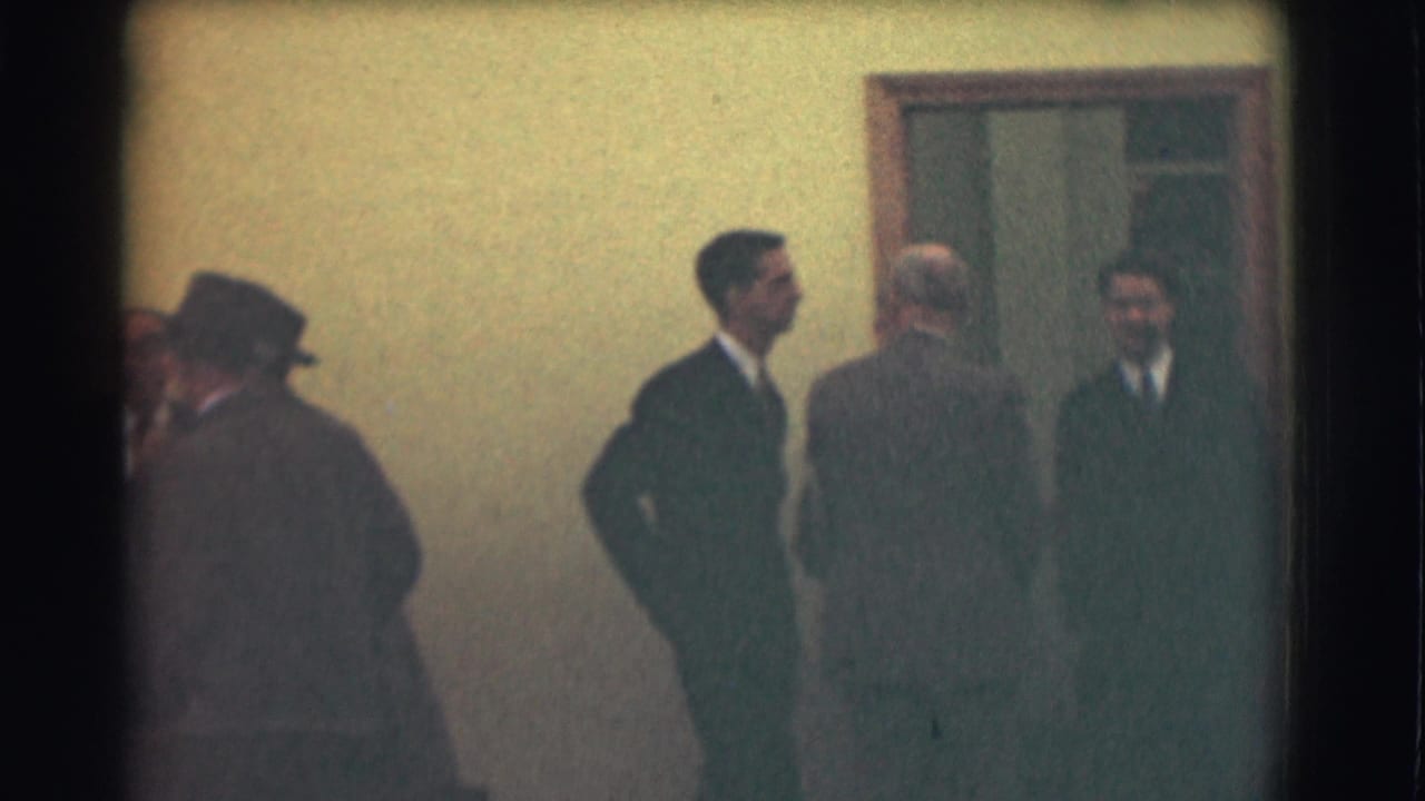 In 1959 Saint Louis footage, several suited men wait and converse outside a doorway, with hats visible at the frame edge.