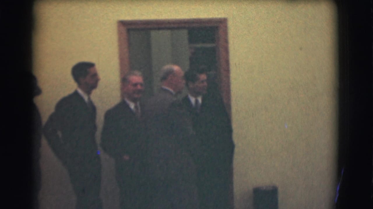 Four formally dressed men stand by a doorway in the 1959 Saint Louis home-movie footage, facing different directions as they wait.
