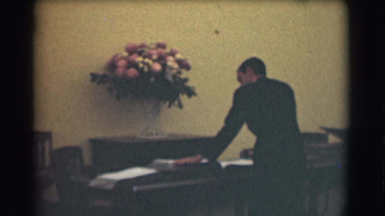 In the 1959 Saint Louis footage, a suited man bends over a desk or table near large volumes and a flower arrangement.