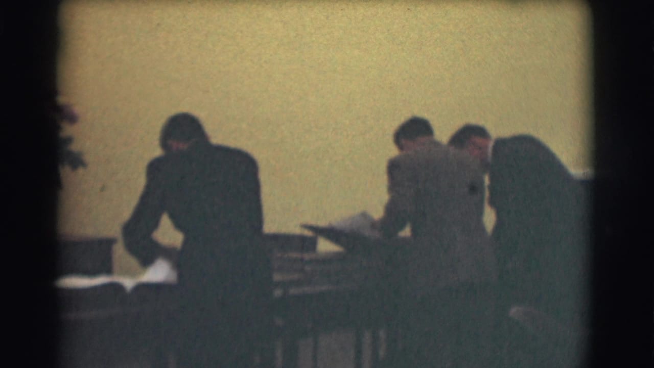 Several men in suits stand around desks and look through large books or papers.