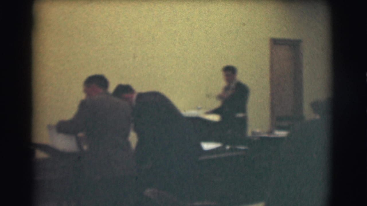 Men in suits work at desks in a mid-century Saint Louis room, with another man standing near the back wall.