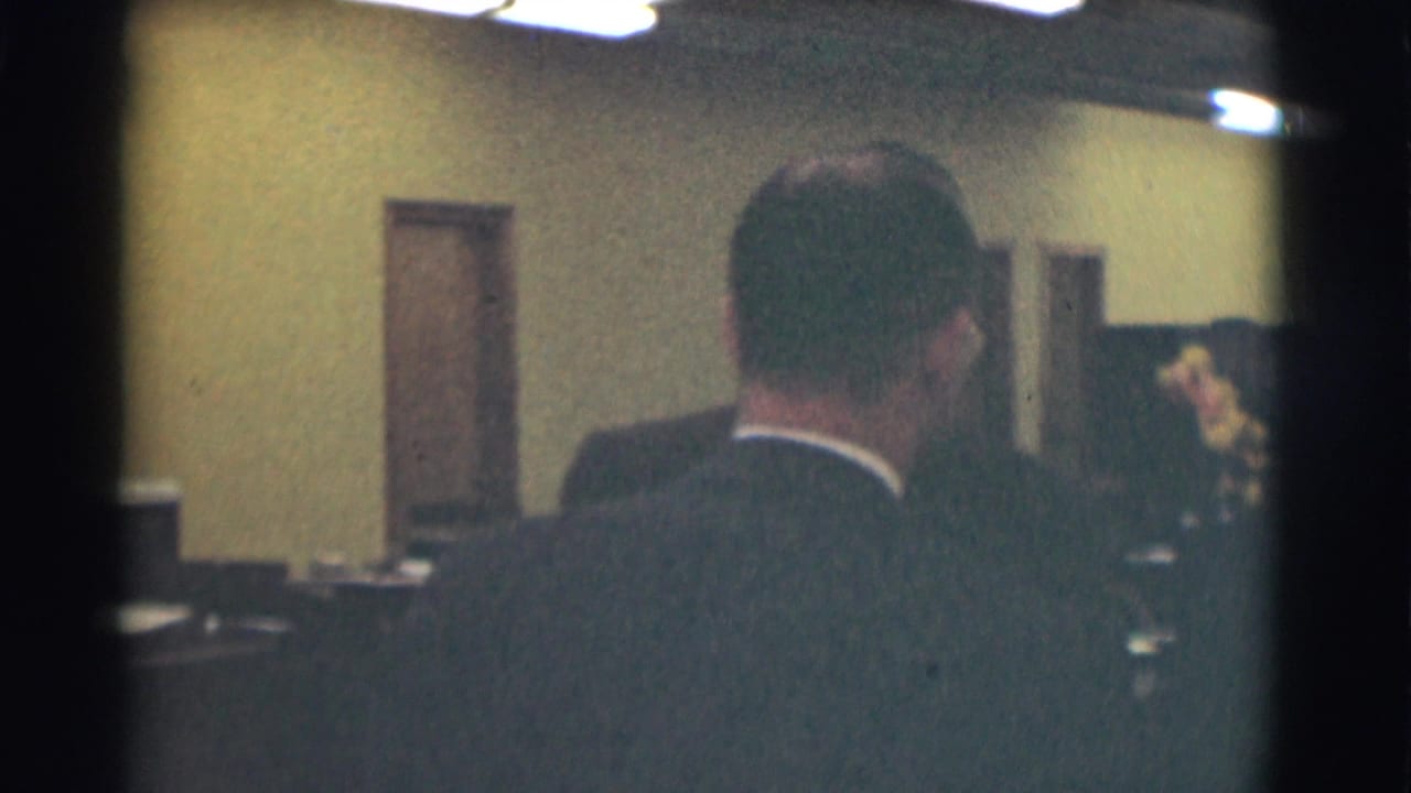 The frame looks past a suited man toward desks, doorways, and another figure in the Saint Louis interior.