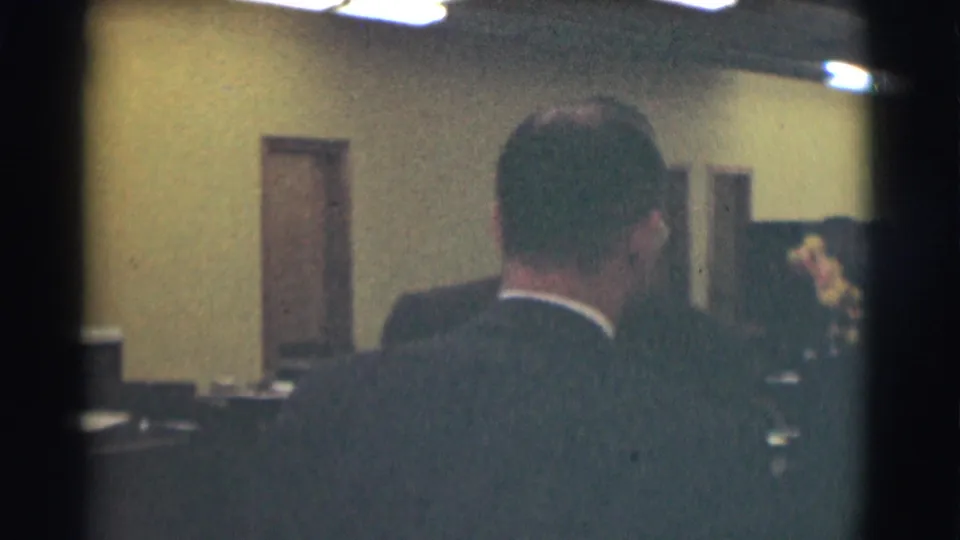 The frame looks past a suited man toward desks, doorways, and another figure in the Saint Louis interior.