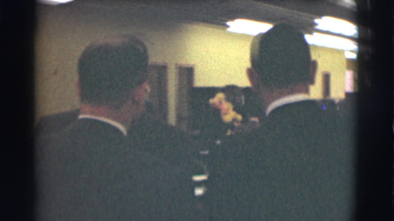 Two men in dark suits seen from behind inside a dim office meeting room.
