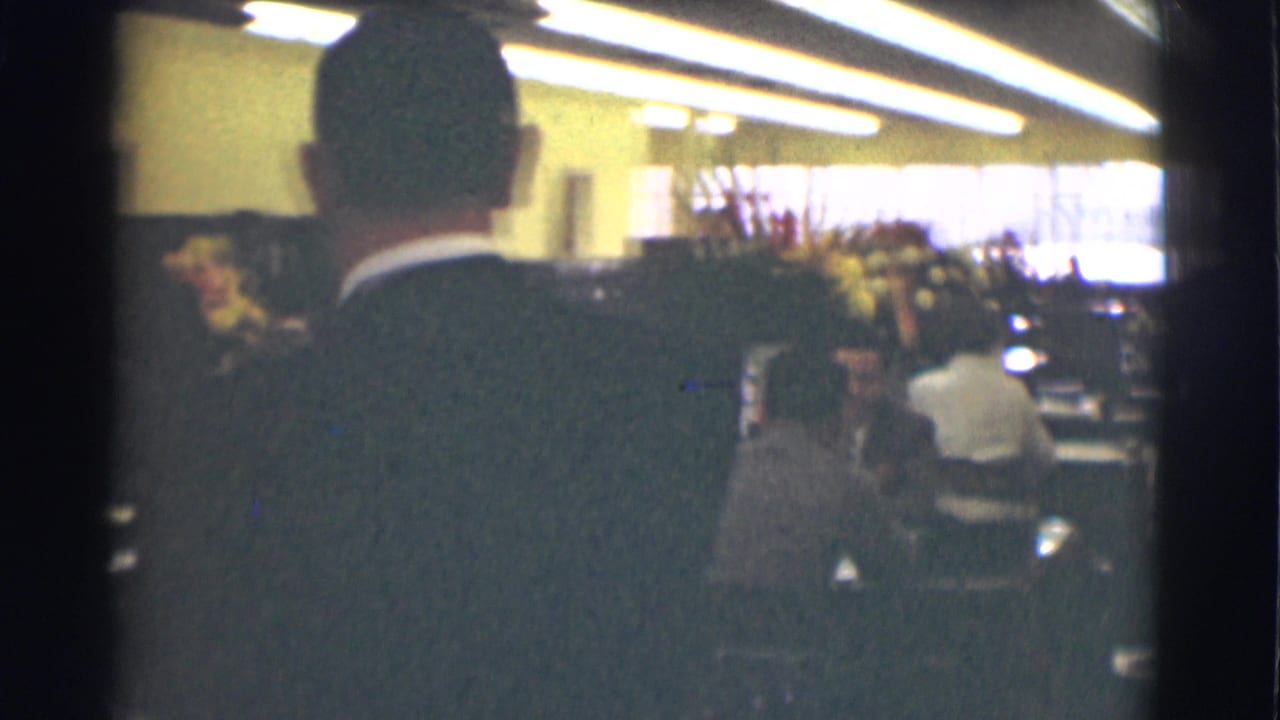 A suited man faces into a 1959 Saint Louis office room with seated adults, tables, floral decor, and bright windows beyond.