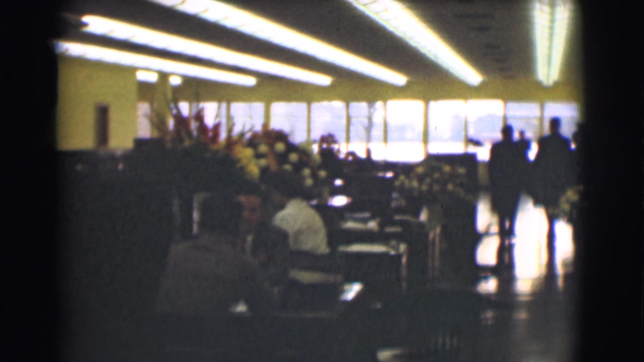 The handheld 8mm view opens to a wider 1959 Saint Louis office interior with meeting tables, chairs, floral arrangements, and adults near windows.