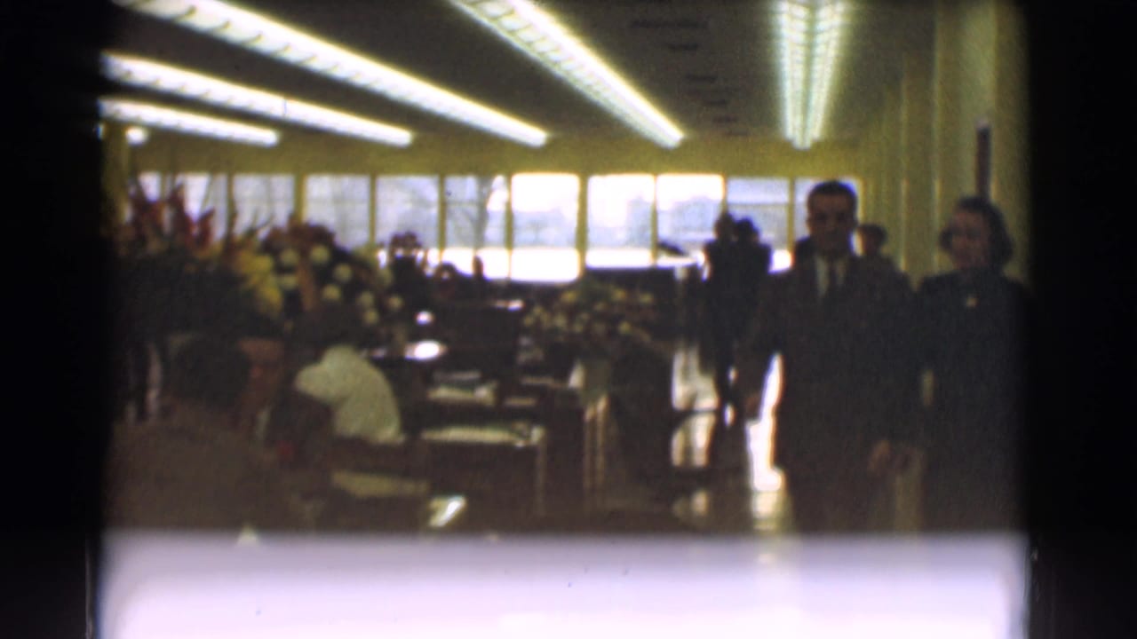 Two suited men move through the 1959 Saint Louis office interior as seated adults, meeting tables, flowers, and bright windows remain visible.