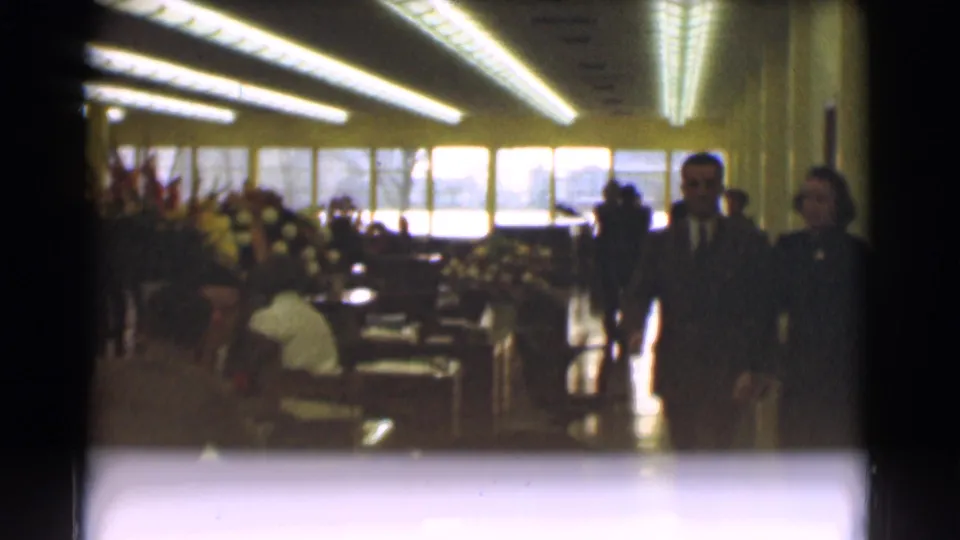 Two suited men move through the 1959 Saint Louis office interior as seated adults, meeting tables, flowers, and bright windows remain visible.