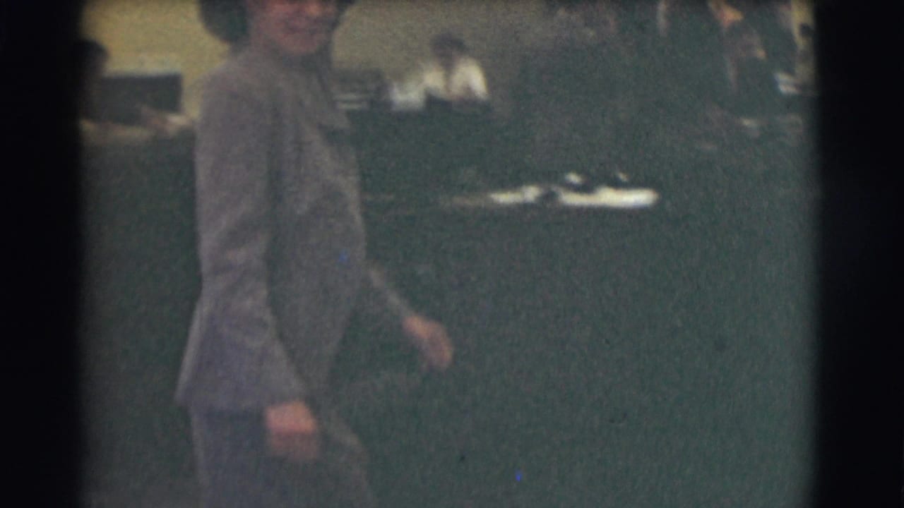 Woman in a gray suit and dark hat walks past desks in a dim office interior.