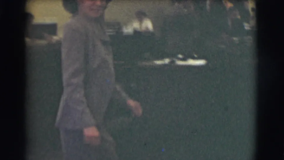 In 1959 Saint Louis footage, a woman in office attire walks through a mid-century workplace with desks behind her.