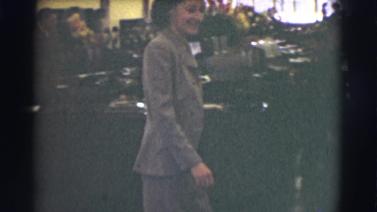 The woman continues through the 1959 Saint Louis office space, smiling slightly as desks and workers fill the background.