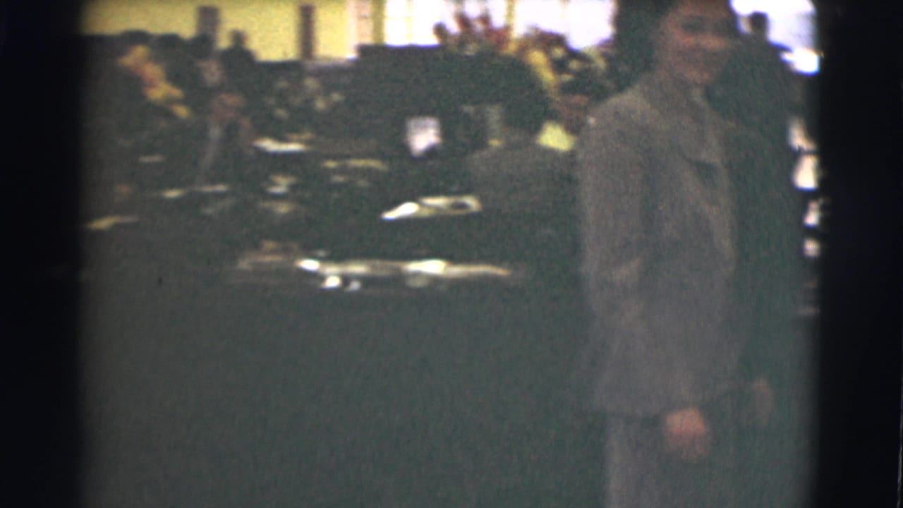 A close view shows the woman in a gray suit near the camera, with office tables, papers, and people behind her.